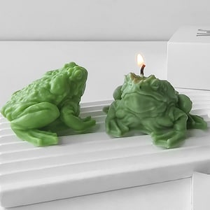 May include: Two green frog-shaped candles, one with a lit wick. The candles are on a white surface.