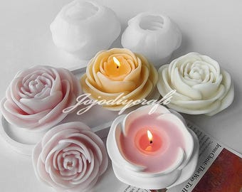 May include: Six white and pink rose-shaped candle holders with a single candle in each. The candles are lit and the flames are visible. The candle holders are arranged in a circle on a white surface.