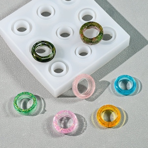 May include: A silicone mould with nine circular cavities for making resin rings. Six rings are shown, including green, blue, pink and yellow rings with glitter and a green ring with dried flowers.