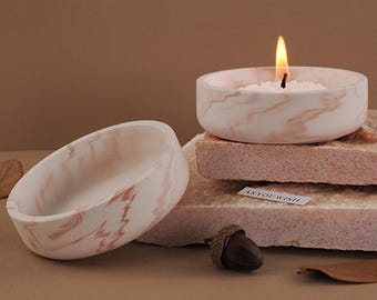 Puede incluir: Dos portavelas de mármol blanco con vetas rosadas. Un portavelas tiene una vela blanca con una mecha encendida en su interior. Los portavelas están sobre una pila de bloques de piedra rosa y blanca. El texto "As You Wish" está impreso en una pequeña etiqueta blanca.
