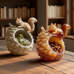 May include: Two decorative containers shaped like pinecones, each featuring a squirrel perched on top. The left container is matte grey and holds a small succulent plant. The right container is translucent amber-coloured resin and holds brown nuts.