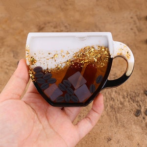 May include: A white ceramic coffee cup filled with resin, featuring gold flakes, coffee beans, and clear ice cubes. The cup is held in a hand.
