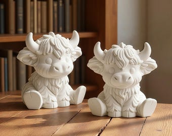 May include: Two matte white ceramic figurines shaped like Highland cattle calves. Each figure features a shaggy coat texture, curved horns, and a seated posture with front legs extended forward. The sculptures are solid white with no colour.