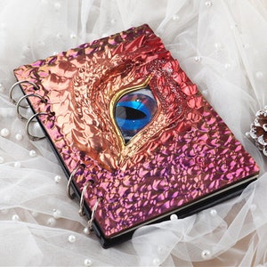 Pu&ograve; includere: Un quaderno a spirale rosa e oro con un designo di occhio di drago. L'occhio &egrave; blu e oro con un motivo di squame di drago rosso e oro intorno.