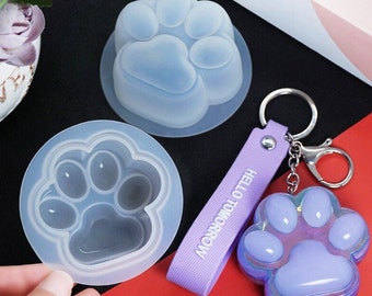 Può includere: Due stampi in silicone trasparente a forma di impronta di zampa, uno più grande dell'altro. Un portachiavi viola con il testo "HELLO TOMORROW" è attaccato a un ciondolo viola a forma di impronta di zampa.
