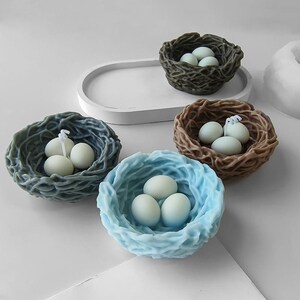 Puede incluir: Cuatro nidos de velas decorativos con tres huevos blancos en cada uno. Los nidos est&aacute;n hechos de un material tejido y son en tonos de marr&oacute;n, azul y gris.