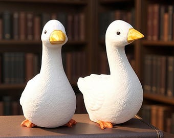 May include: Two white goose ornaments with yellow beaks and orange feet are displayed on a brown surface. The geese have a textured surface and are positioned in front of a blurred background of bookshelves. The ornaments are decorative.