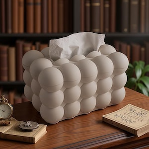 Può includere: Un porta fazzoletti beige a forma di bolle, con fazzoletti bianchi che spuntano. È appoggiato su un tavolo di legno, accanto a un piccolo orologio da tasca e a un libro d'epoca. Sullo sfondo, una libreria piena di libri.
