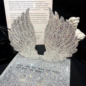 Op de afbeelding: Een paar zilveren glitter-engelenvleugels met een wit gipsen buste van een mannenhoofd op de achtergrond. De vleugels staan op een zilveren glitterbasis met de tekst "LOVE LOOK NOT WITH THE EYES BUT WITH THE MIND".