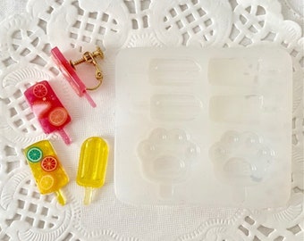 May include: A collection of handmade resin popsicle earrings and a mold. The earrings are in the shape of popsicles with fruit slices. The mold is white and has popsicle and paw print shapes.