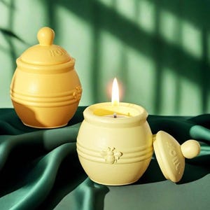 Puede incluir: Dos tarros de velas de cerámica amarilla con tapas con forma de tarros de miel. El tarro de vela de la derecha tiene una vela encendida dentro y un diseño de abeja en la parte delantera. El tarro de la izquierda tiene la palabra "HONEY" escrita en la tapa.