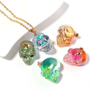 K&ouml;nnte beinhalten: F&uuml;nf regenbogenfarbene Glassch&auml;del-Anh&auml;nger in verschiedenen Farben, darunter Gr&uuml;n, Pink und Orange. Die Sch&auml;del sind alle detailliert mit Augenh&ouml;hlen, einer Nase und einem Mund. Einer der Sch&auml;del ist an einer goldenen Kette befestigt.