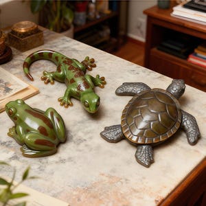 Puede incluir: Tres figuras de animales de cerámica: una rana verde con manchas marrones, un lagarto verde con marcas marrones y una tortuga de color bronce. Las figuras se exhiben sobre una superficie con diseño de mármol.