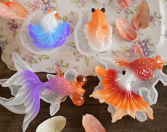 May include: A collection of colorful, translucent goldfish figurines in various poses. The fish are in shades of orange, purple, blue, and white, with detailed fin and tail designs. Some are set in clear molds, and scattered petals add to the decorative arrangement.