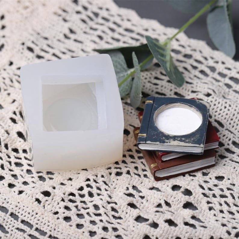 May include: A white silicone mold shaped like a book with a square opening in the center. Next to the mold is a stack of three miniature books with a blue cover and gold accents.