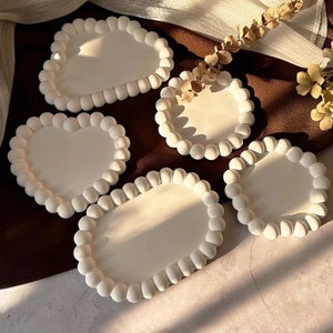 Peut inclure: Quatre sous-verres en céramique blanche avec un design texturé en relief, ressemblant à des bulles, autour des bords. Les sous-verres sont en forme de cœur, d'ovale et de deux cercles.