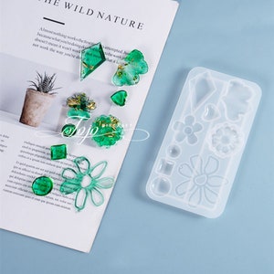 Peut inclure: Un moule en silicone transparent avec 12 formes différentes, y compris des fleurs, des cœurs et des formes géométriques. Le moule est sur une surface bleu clair. Il y a plusieurs pièces de résine verte à côté du moule.