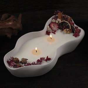 Puede incluir: Portavelas de cerámica blanca con forma de ola con dos velas encendidas y detalles de flores y frutas secas.