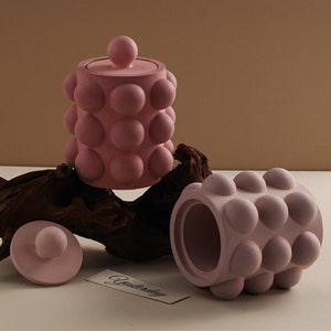 Könnte beinhalten: Zwei rosa Keramikbehälter mit einem strukturierten, blasenartigen Design. Die Behälter haben Deckel mit einem runden Knopf oben. Die Behälter stehen auf einem Stück Treibholz.