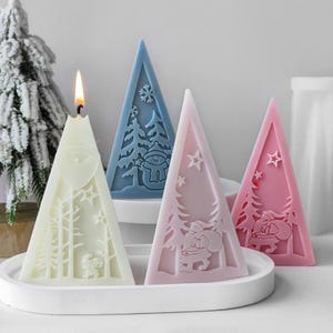 May include: Three triangular Christmas tree candles in white, blue and pink. Each candle features a different winter scene with a snowman, a reindeer and a tree.