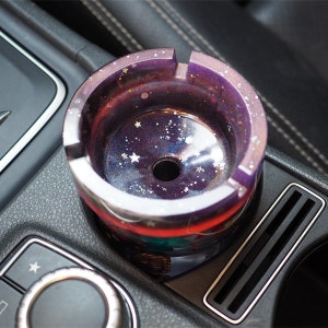 May include: A purple and white galaxy-themed car ashtray with a star pattern and glitter. The ashtray is sitting in a car's cup holder.