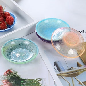 May include: Four small, round, clear resin dishes with glitter embedded in the resin. The dishes are in various colors, including blue, green, pink, and purple.