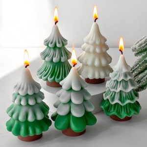 Puede incluir: Un conjunto de velas con forma de árbol de Navidad en varios tonos de verde y blanco. Cada vela tiene una mecha encendida, creando un brillo cálido. Las velas están dispuestas sobre una superficie blanca, evocando un tema festivo.