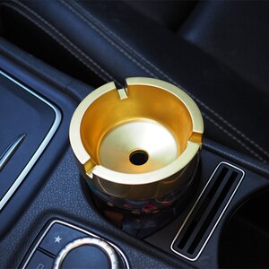 May include: A gold-colored car ashtray with a black base. The ashtray is sitting in a car's cup holder.