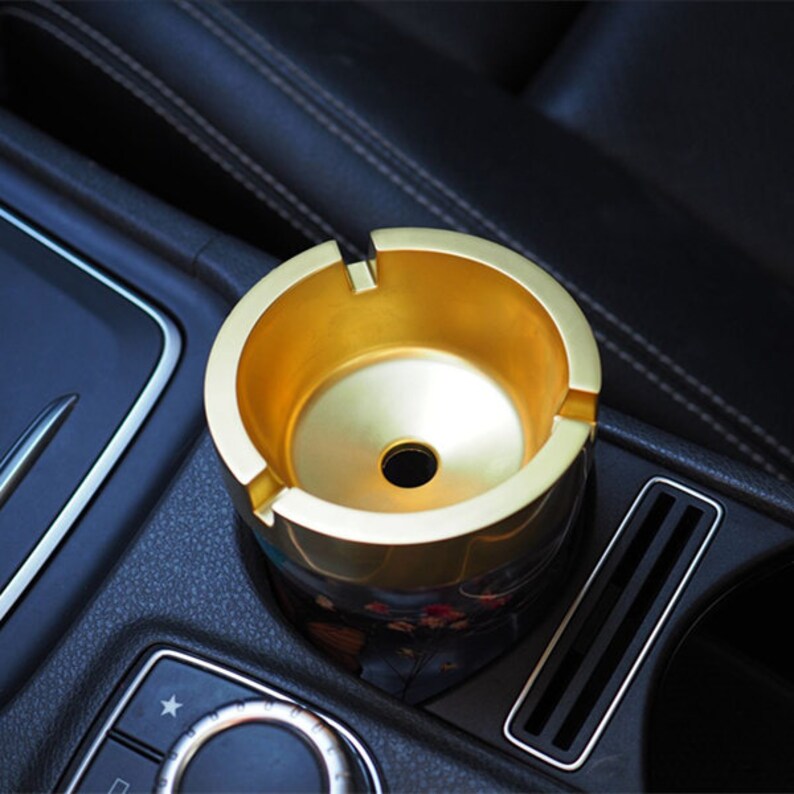 May include: A gold-colored car ashtray with a black base. The ashtray is sitting in a car's cup holder.