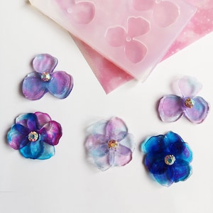 May include: Six resin flower embellishments with iridescent glitter and rhinestones in shades of blue, purple, and pink. The flowers are arranged on a white surface.
