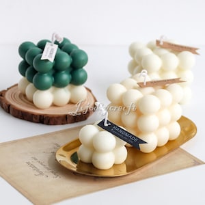 May include: Three handmade bubble candles in green, white, and white. The candles are on wooden and gold metal bases. The candles have small tags that say "Handmade" on them.