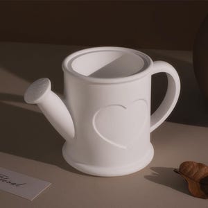 Puede incluir: Una regadera de cerámica blanca con un corazón en relieve en la parte delantera. La regadera tiene un pico con pequeños agujeros y un asa curvada. El diseño es simple y elegante, adecuado para uso en interiores o exteriores.