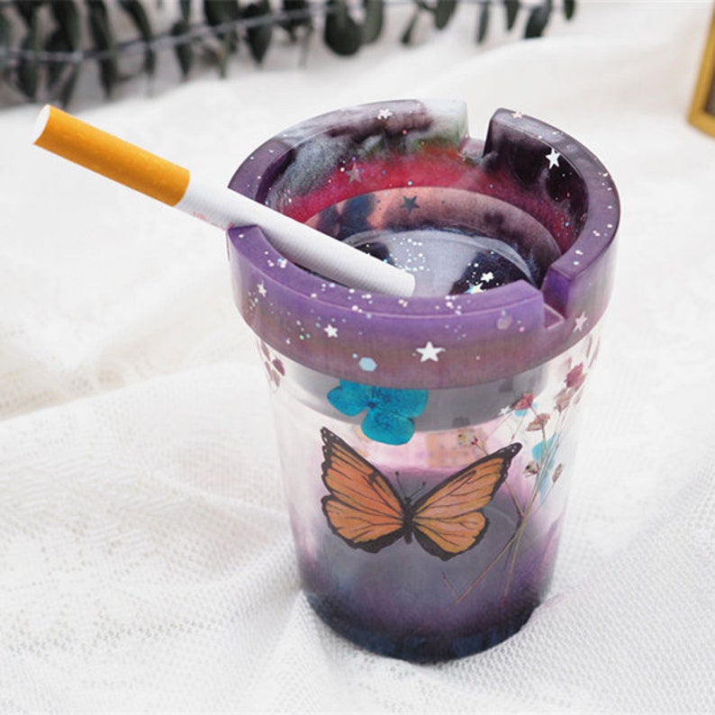 May include: Purple and clear glass ashtray with a galaxy design and a butterfly on the side. The ashtray has a white rim with stars and a white cigarette inside.