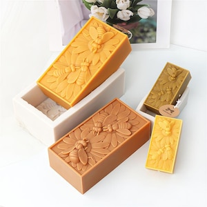 May include: Three rectangular beeswax candles with a raised design of bees and flowers. The candles are in different sizes and are arranged in a row from largest to smallest.