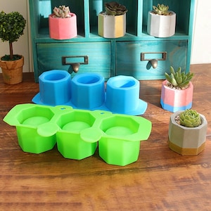 May include: A selection of small, geometric planters in shades of pink, blue, and grey, each containing a succulent plant. Also shown are green and blue silicone moulds for creating similar planters. The planters are displayed on a wooden surface.