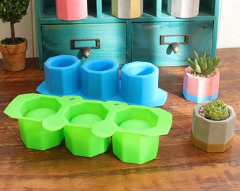 May include: A collection of small, geometric planters in various colors, including pink, blue, and gray, each holding a succulent plant. Also shown are green and blue silicone molds for creating similar planters. The planters are displayed on a wooden surface.