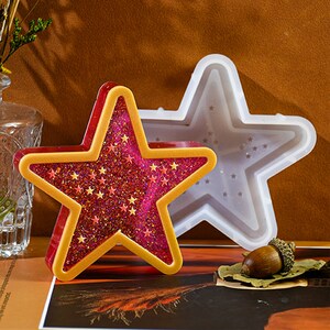 Puede incluir: Un molde de silicona de dos piezas para hacer manualidades de resina. El molde tiene forma de estrella con un contorno dorado y un interior de purpurina rosa. El molde se muestra con una estrella de resina terminada.