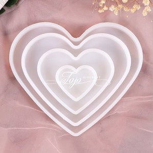 Puede incluir: Tres moldes de silicona en forma de corazón de diferentes tamaños, anidados uno dentro del otro. Los moldes están sobre un fondo rosa y blanco.