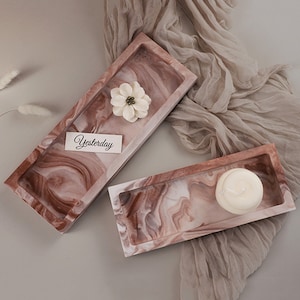 Può includere: Due vassoi rettangolari con un designo marmoreo in tonalità di marrone, rosa e bianco. I vassoi sono vuoti, ma uno contiene un fiore bianco e la parola "Yesterday" scritta su un biglietto all'interno.