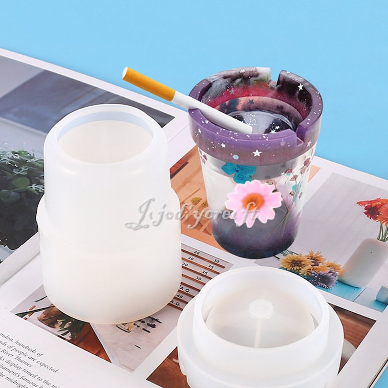 May include: Two silicone molds for making a resin ashtray. The mold is white and has a cylindrical shape. The ashtray is purple with a galaxy design and white stars. A cigarette is resting on the rim of the ashtray.
