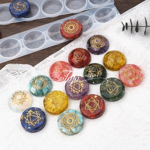 Peut inclure: Ensemble de 12 pierres de chakra avec des motifs géométriques dorés. Chaque pierre est d'une couleur différente et présente un symbole de chakra unique. Les pierres sont disposées en cercle sur une nappe en dentelle blanche.