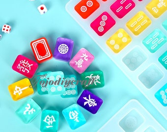 Puede incluir: Un molde de silicona transparente con 16 compartimentos individuales con forma de fichas de mahjong. El molde está sobre una superficie azul con 12 fichas de mahjong de colores, cada una con un símbolo y un color diferente.