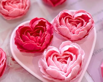 May include: Four pink and red heart-shaped flower arrangements on a pink heart-shaped plate. The flowers are made of soap and have a delicate, layered appearance.