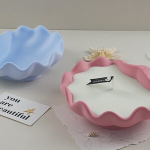 May include: Two decorative shell-shaped candle holders. One is light blue and the other is pink. The pink holder has a white candle inside with a small flag that says "forever".