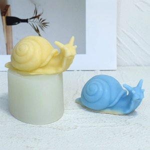 Peut inclure: Deux bougies en forme d'escargot, l'une jaune et l'autre bleue, sur une surface blanche. L'escargot jaune est sur un piédestal blanc.
