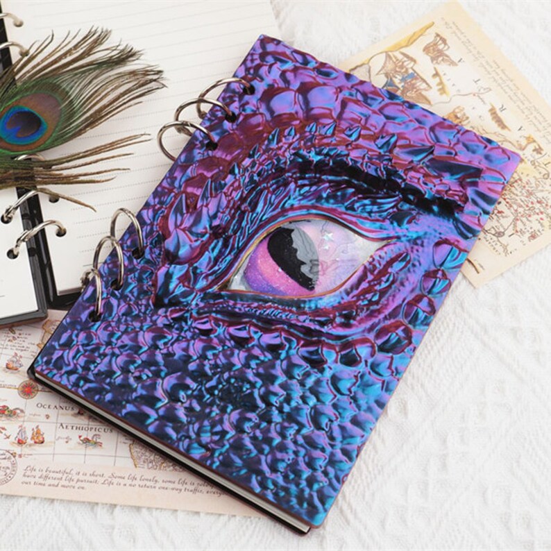 Pu&ograve; includere: Un quaderno a spirale con una copertina iridescente viola e blu che presenta un occhio di drago con un'iride rosa e bianca. Il quaderno &egrave; su una superficie bianca con una mappa e una piuma di pavone.