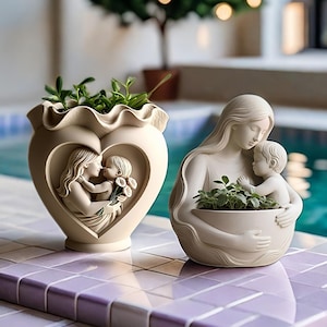 May include: Two cream-colored resin planters featuring sculpted figures of a parent and child. One planter is heart-shaped with a ruffled rim, while the other depicts a figure cradling a child. Both contain small green succulent plants.