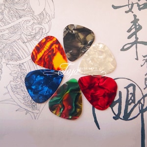 Puede incluir: Seis púas de guitarra en una variedad de colores y patrones, incluyendo rojo, azul, verde, blanco, negro y naranja. Las púas están dispuestas en un círculo sobre una superficie blanca.