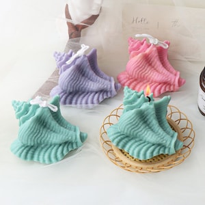 Puede incluir: Tres velas decorativas con forma de concha marina en morado, rosa y verde azulado. Las velas están sobre una superficie blanca con una cesta de mimbre debajo de la vela verde azulada.