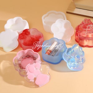 Può includere: Sei scatole di resina a forma di impronta di zampa con coperchi in vari colori, tra cui rosa, blu, bianco e rosso. Le scatole sono riempite con piccoli oggetti, come un braccialetto, rossetto e altri gingilli.